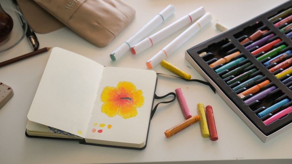A sketchbook with a vibrant flower drawing, surrounded by colorful art supplies on a desk.
