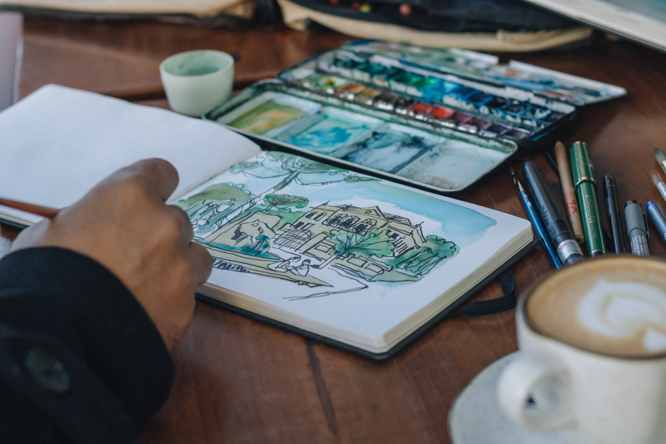 An artist sketching a landscape in a notebook with watercolors, coffee nearby.